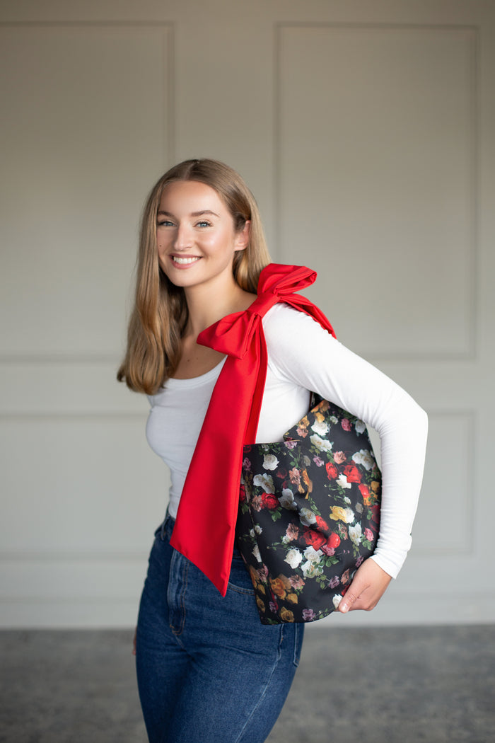 Darling Rose Bow Tote Bag front view, featuring colorful floral pattern on black fabric and bold red bow detail.