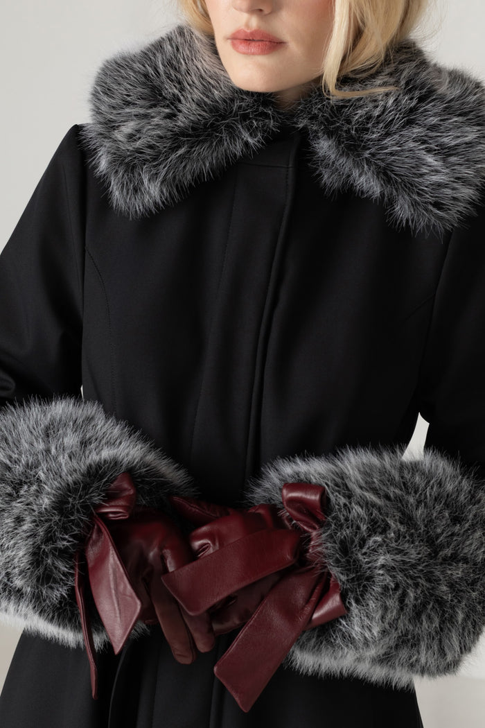 A close-up of a model wearing a black coat with a large gray fur collar and matching gray fur cuffs, accessorized with dark burgundy leather gloves tied with wide ribbons.