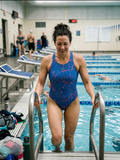 Woman in a blue swimsuit stepping out of an indoor pool.