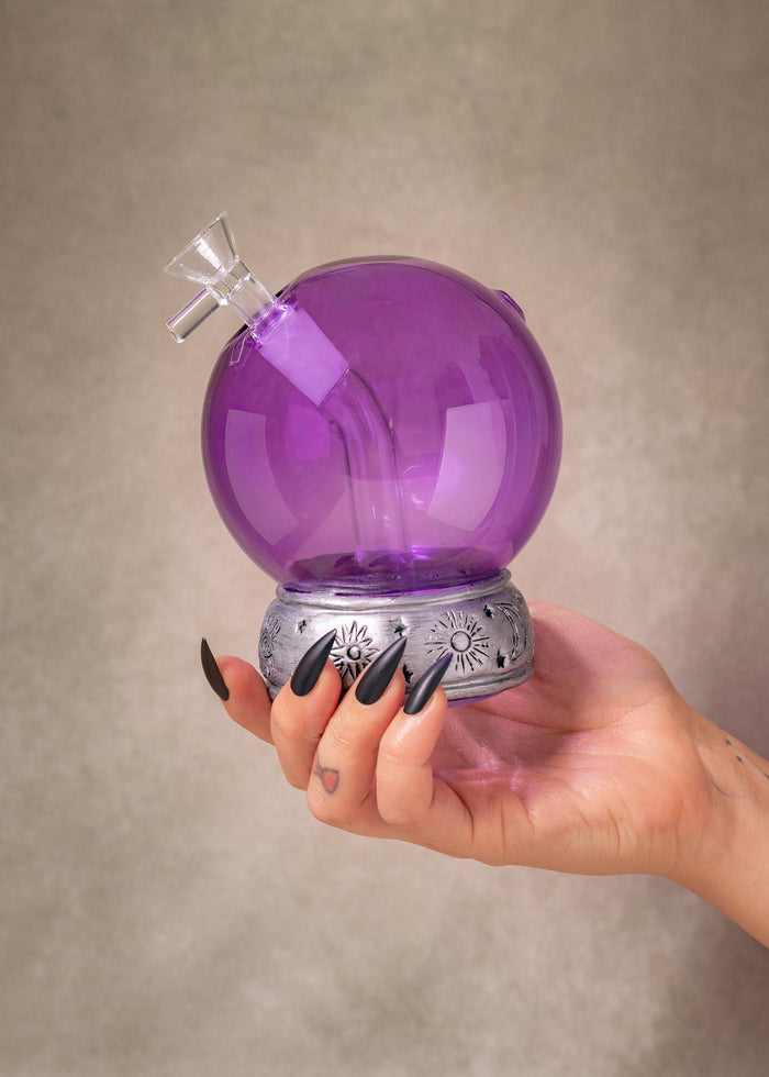 A hand with long black fingernails holds a purple crystal ball glass water pipe with a silver base against a textured beige background.