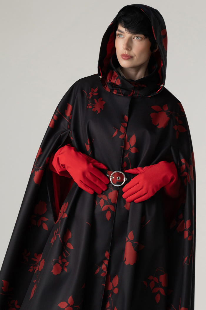 A model wearing a black hooded cape with a red floral pattern, matching red gloves, and a red belt with a silver buckle, photographed against a neutral light gray background.