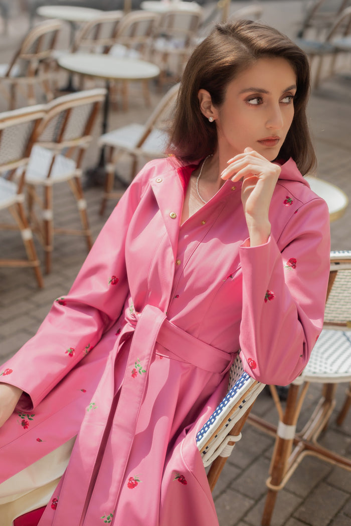 A woman wearing RainSisters pink hooded raincoat Raspberry Sorbet with rose print, sitting on a wicker chair in an outdoor cafe setting.